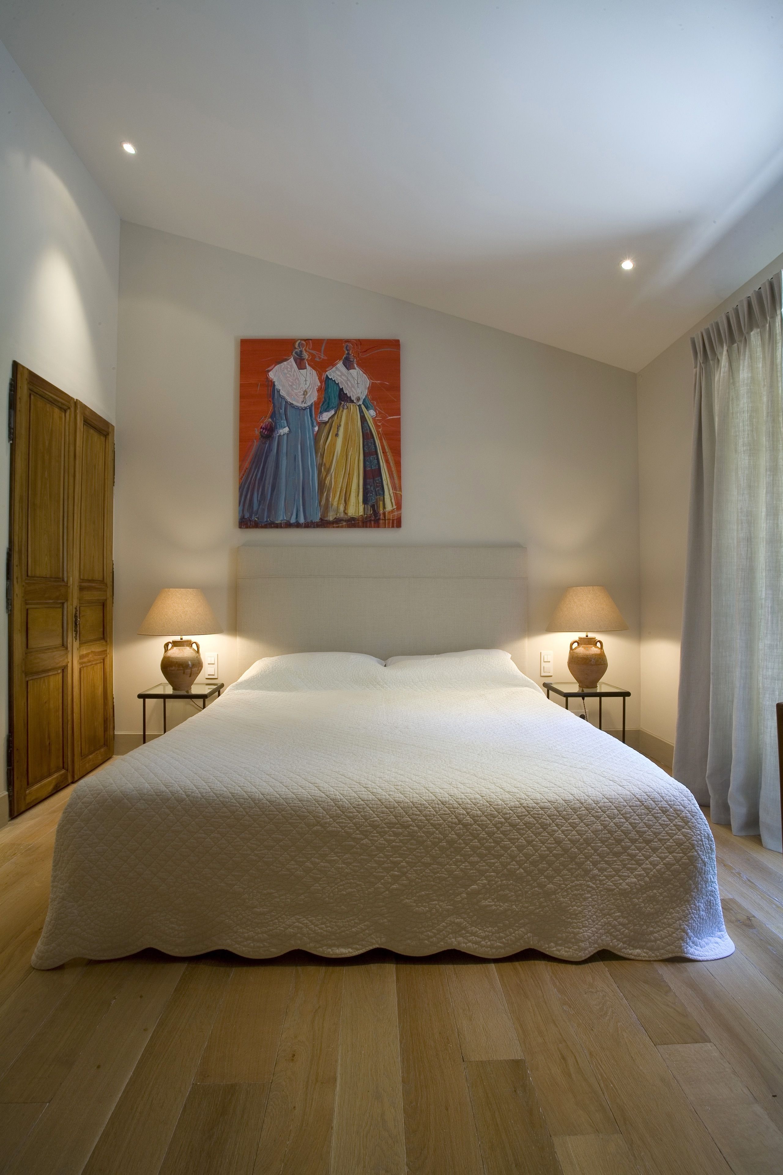 Un grand lit recouvert d’une courtepointe blanche, deux lampes de chevet, un tableau représentant deux personnages accroché au mur et une armoire en bois.