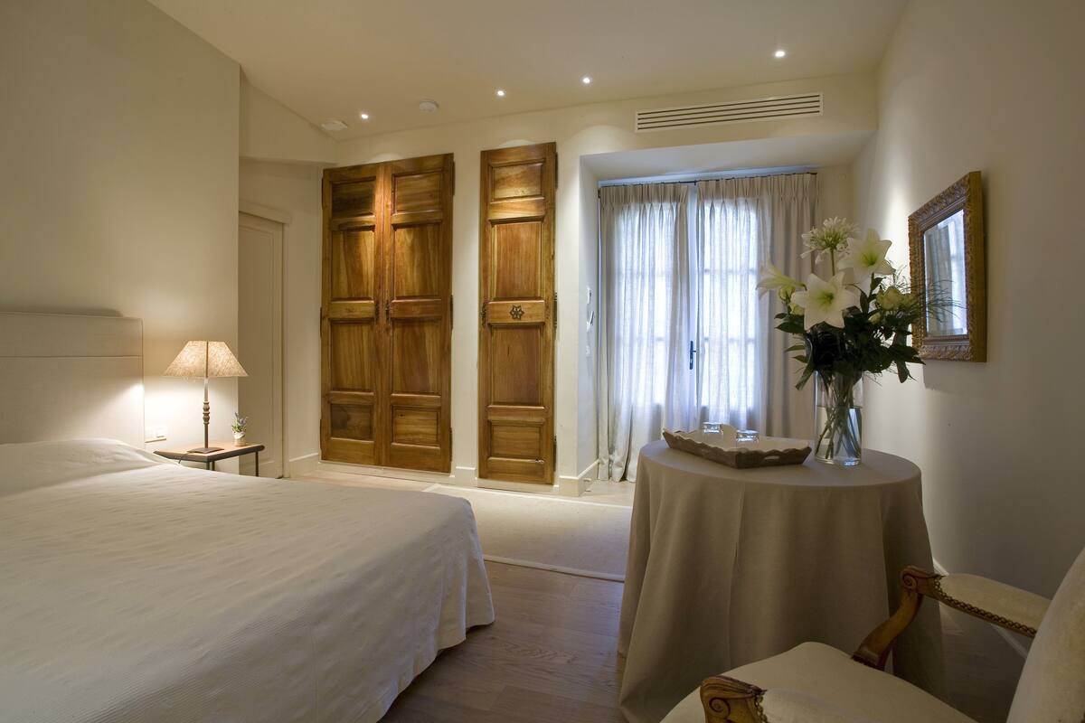 Une chambre d’hôtel avec un grand lit, une petite table sur laquelle est posé un vase de fleurs, une chaise et une grande fenêtre avec des rideaux.