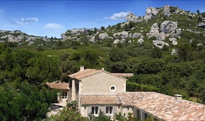 A traditional stone building with a tiled roof, surrounded by lush greenery and rocky hills. - Benvengudo (Les Baux-de-Provence)