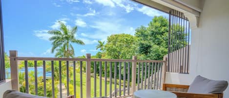 A balcony with a view of a tropical landscape, including palm trees and a body of water. There is a wicker chair with a cushion and a small table.