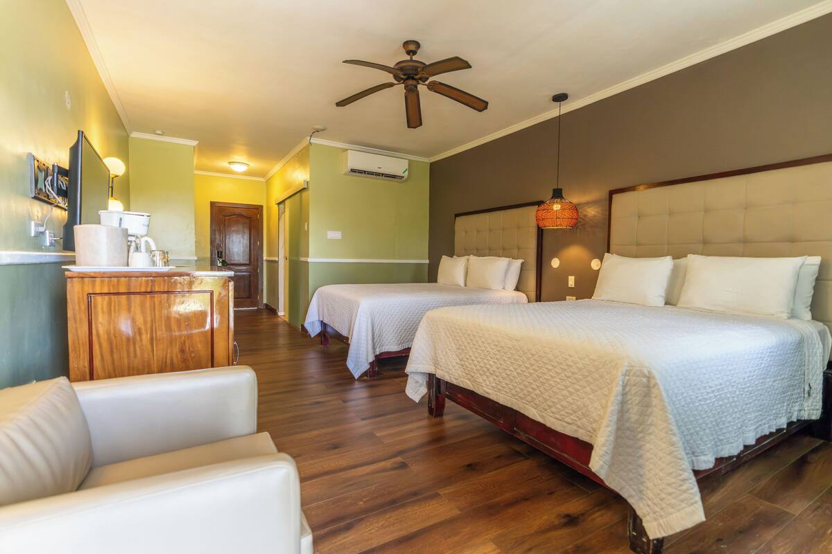 Une chambre d’hôtel avec deux lits, une commode en bois, un ventilateur de plafond et un couloir menant à une autre pièce.