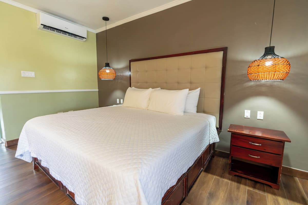 Un lit bien fait, recouvert d’une couette blanche, une table de chevet en bois avec des tiroirs, et deux lampes suspendues avec des abat-jour orange.