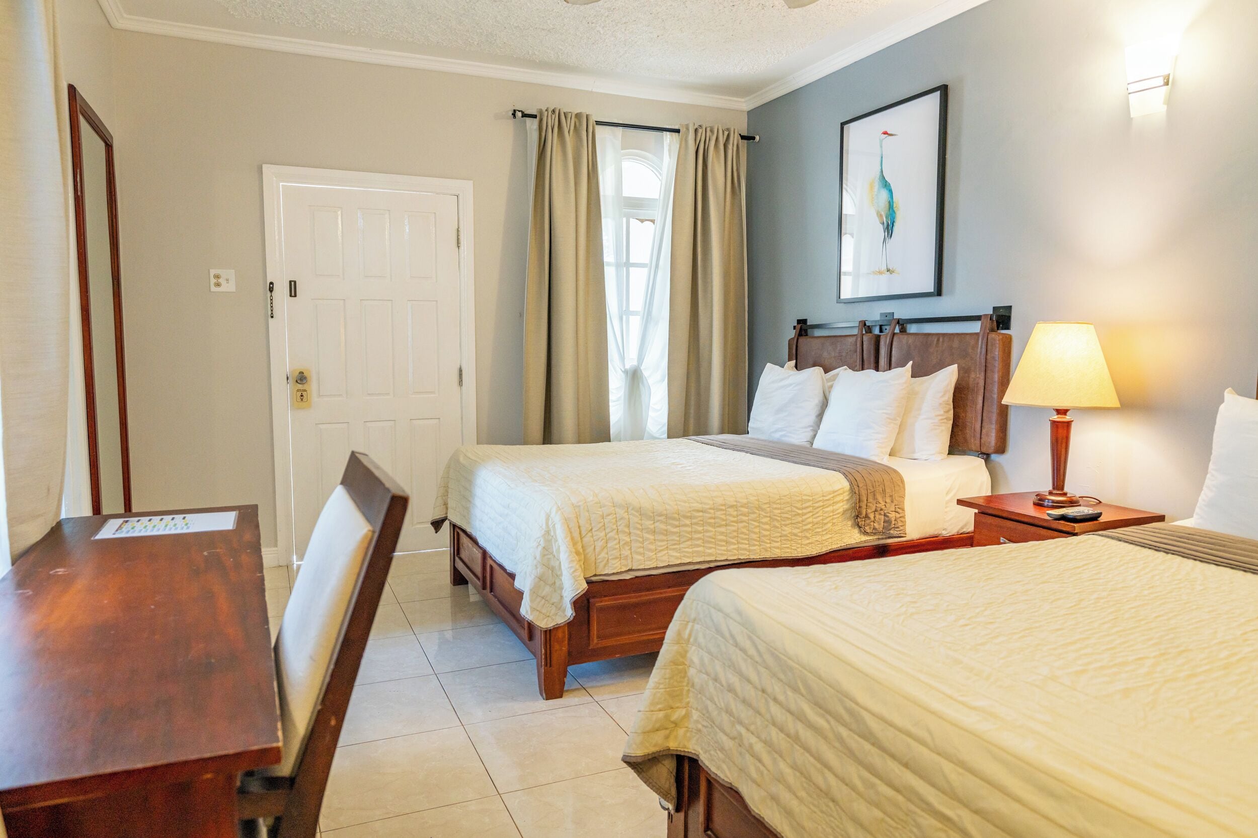 A hotel room with two beds, a desk, a chair, a lamp, and a door.
