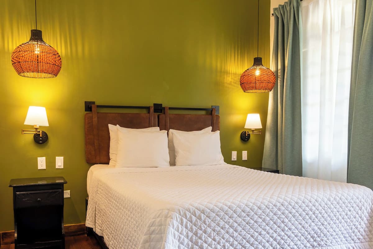 Une chambre d’hôtel avec un lit, deux lampes fixées au mur, une table de chevet et une fenêtre avec des rideaux.
