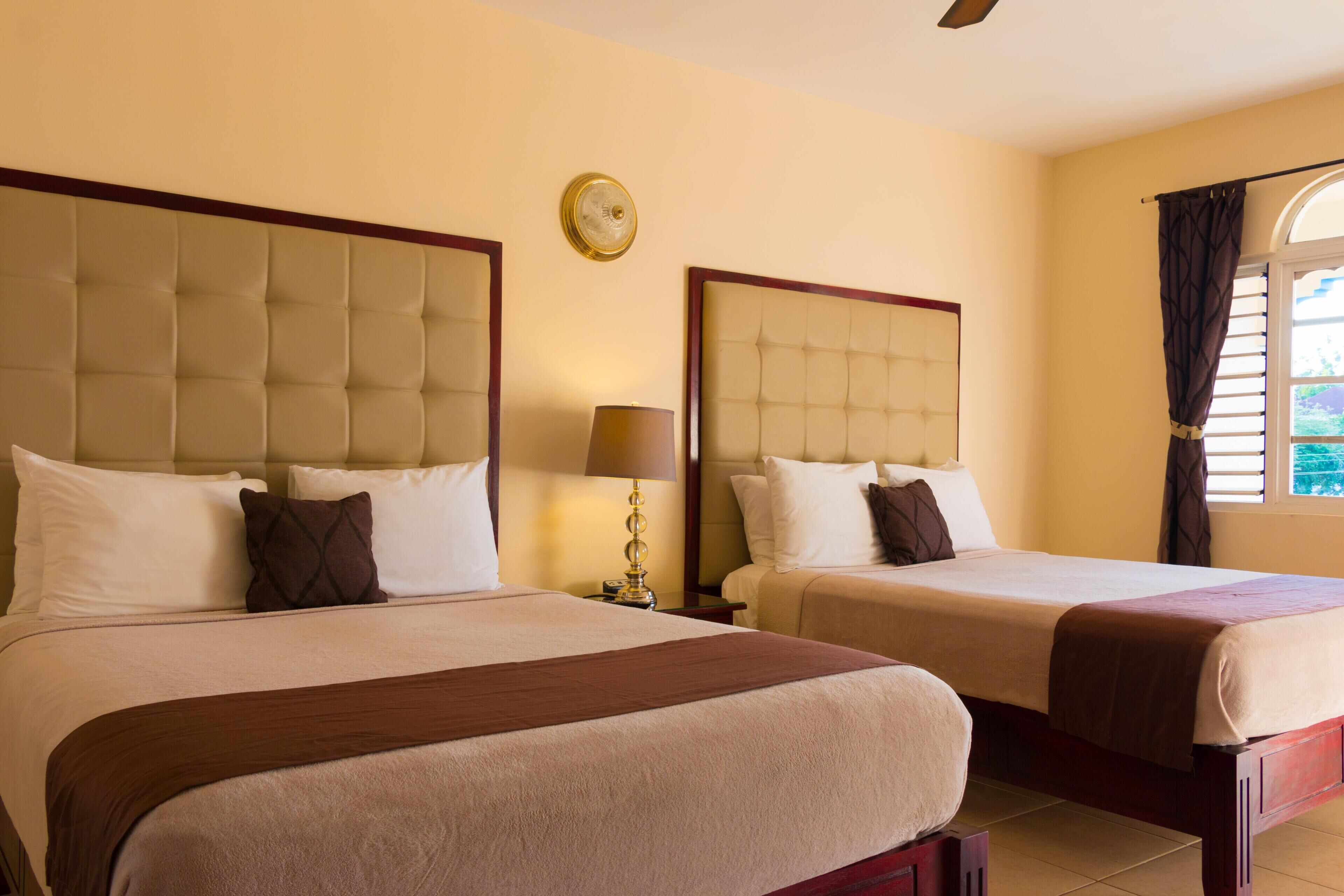 Habitación de hotel con dos camas, una mesita de noche con lámpara, un reloj en la pared y una ventana con cortinas.