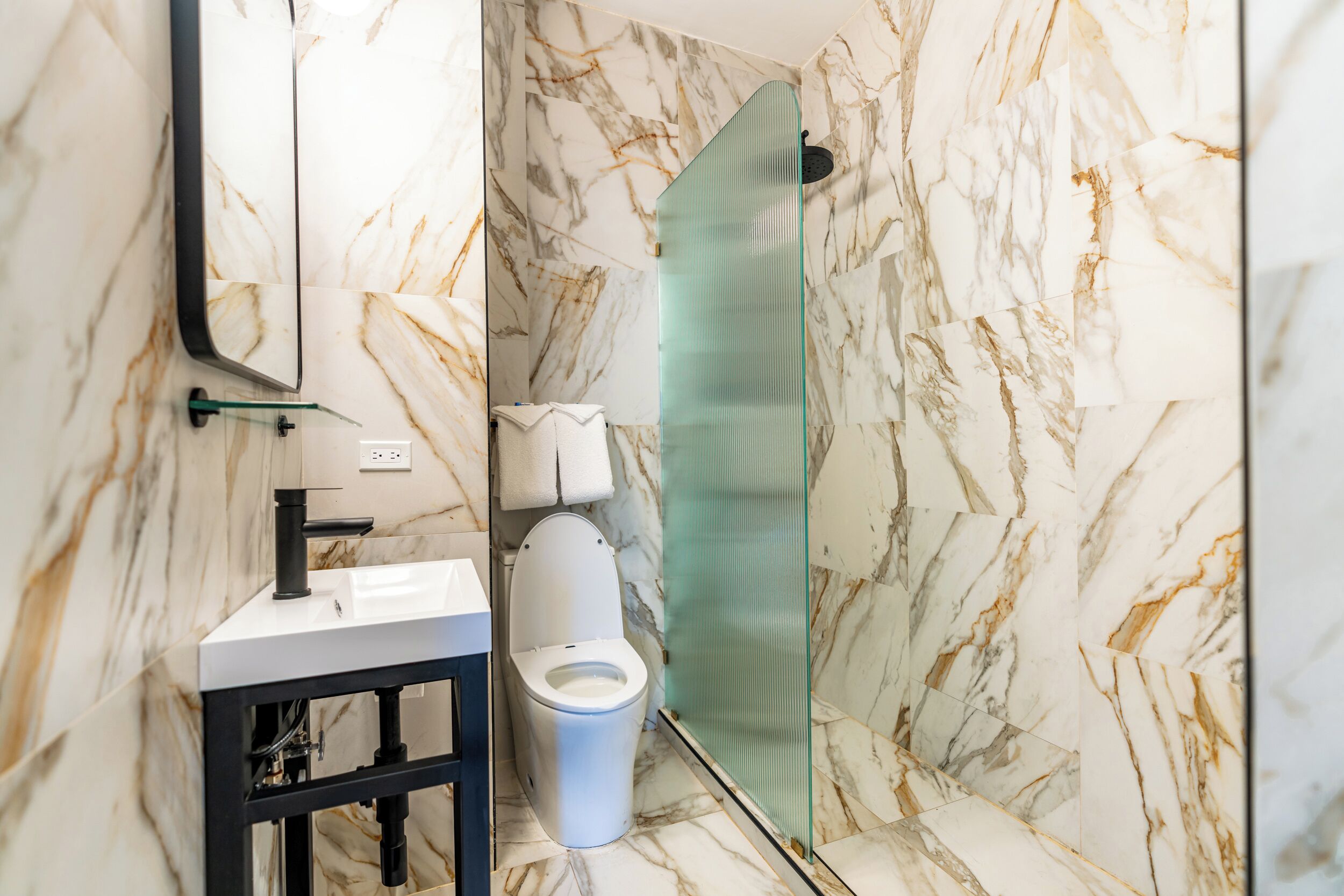 Un baño con pared de mármol, cabina de ducha de cristal, inodoro blanco y lavamanos con marco negro.
