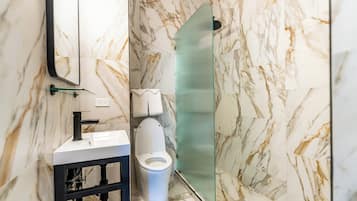 Un baño con pared de mármol, cabina de ducha de cristal, inodoro blanco y lavamanos con marco negro.