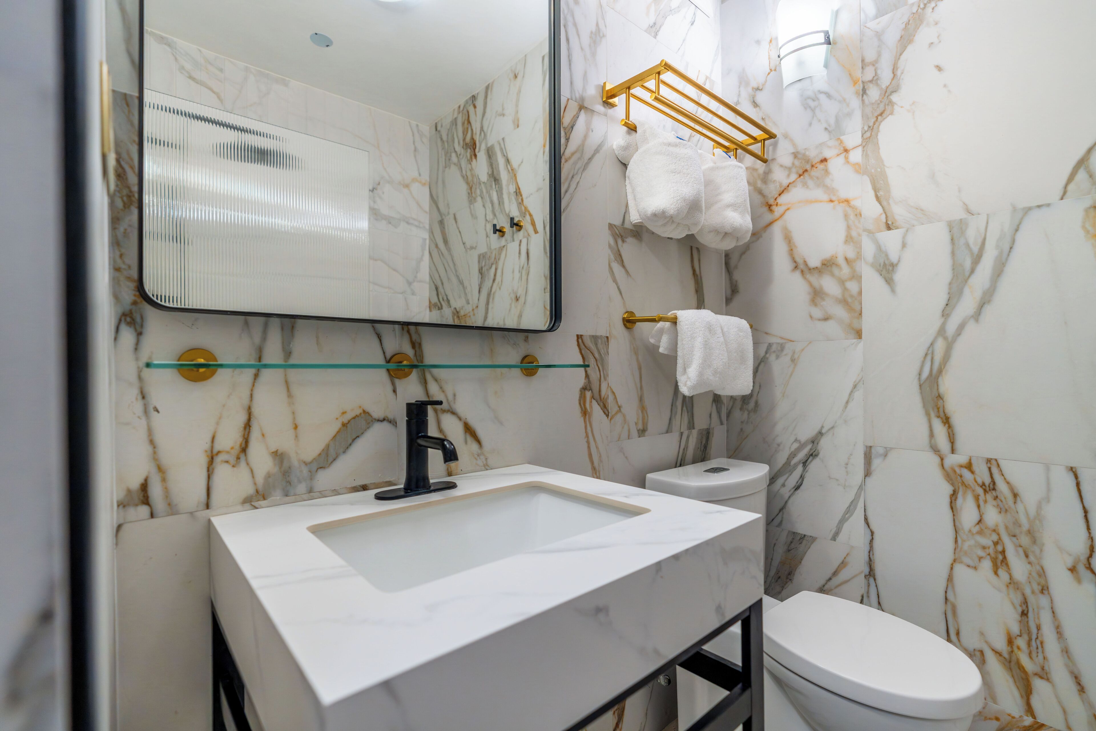 A bathroom with a marble wall, a white sink, a toilet, and a glass shelf above the sink.
