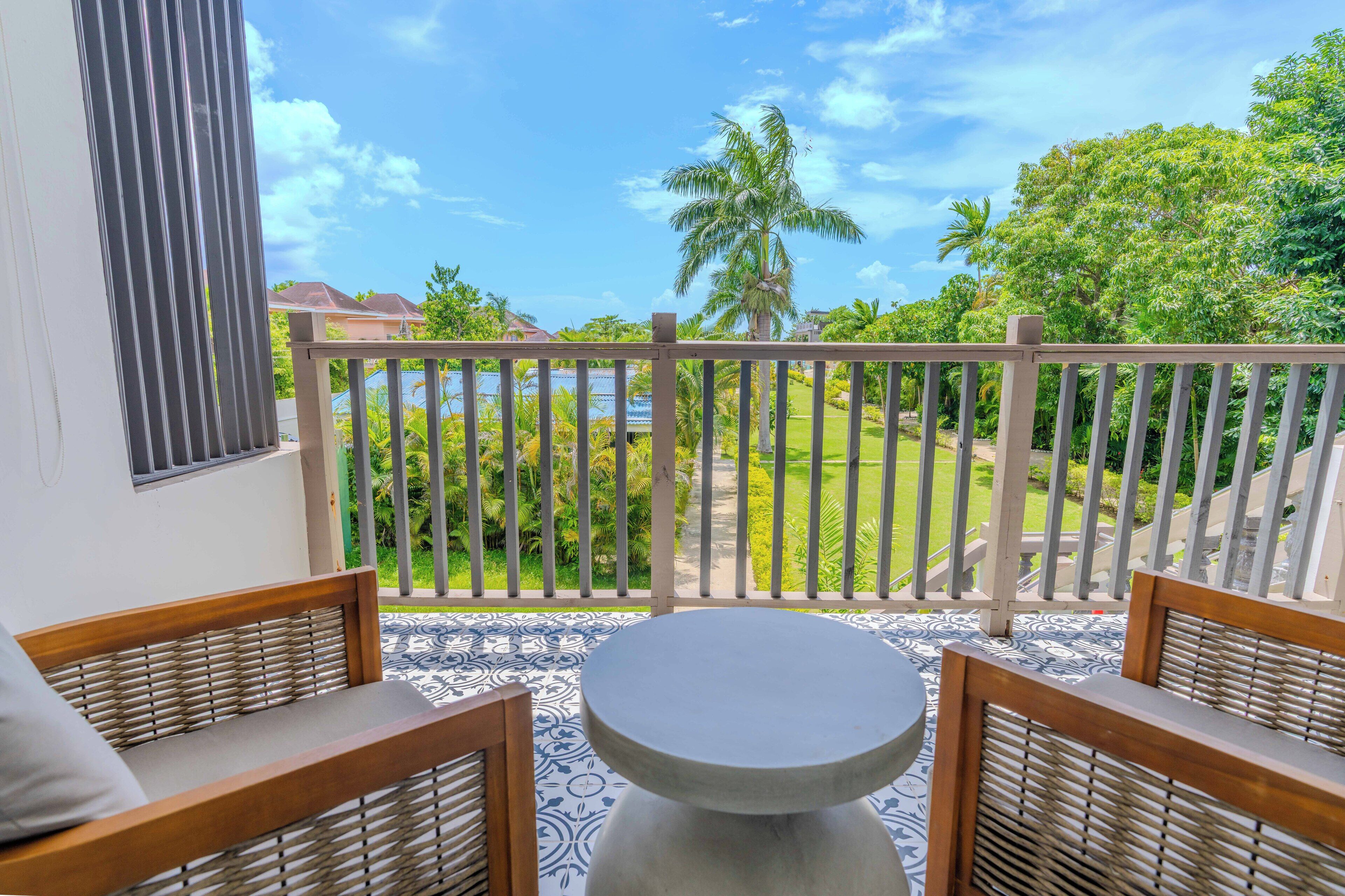 A balcony with a view of a lush garden and a body of water. There are two wooden chairs and a round table.
