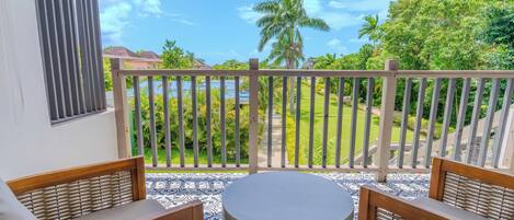 A balcony with a view of a lush garden and a body of water. There are two wooden chairs and a round table.
