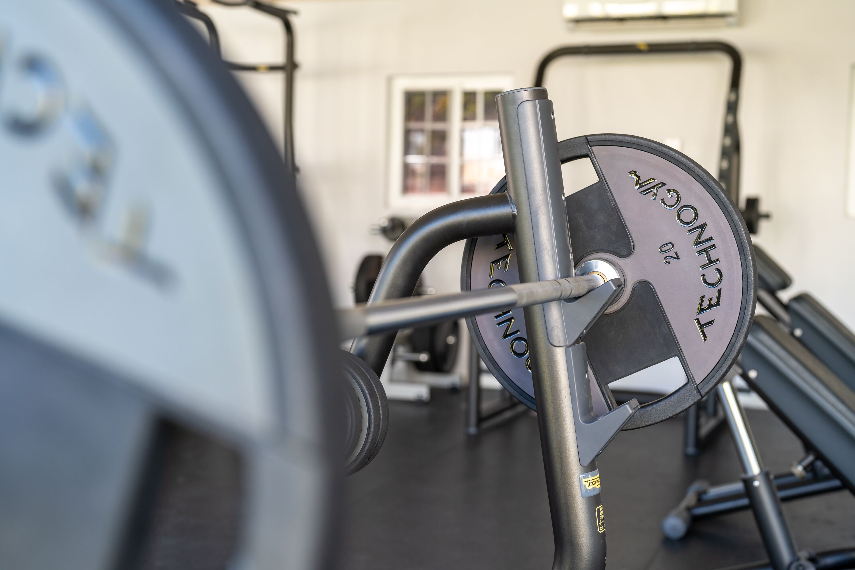 Un banc de musculation avec une barre chargée de poids, placé dans une salle de sport. On aperçoit une fenêtre et d’autres appareils d’exercice à l’arrière-plan.