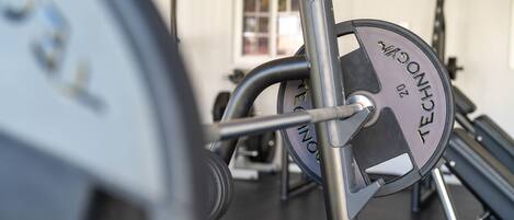 Un banc de musculation avec une barre chargée de poids, placé dans une salle de sport. On aperçoit une fenêtre et d’autres appareils d’exercice à l’arrière-plan.