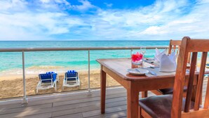 Una terraza de madera con una mesa dispuesta para dos, con vista a una playa que tiene dos sillas reclinables azules y una orilla arenosa.