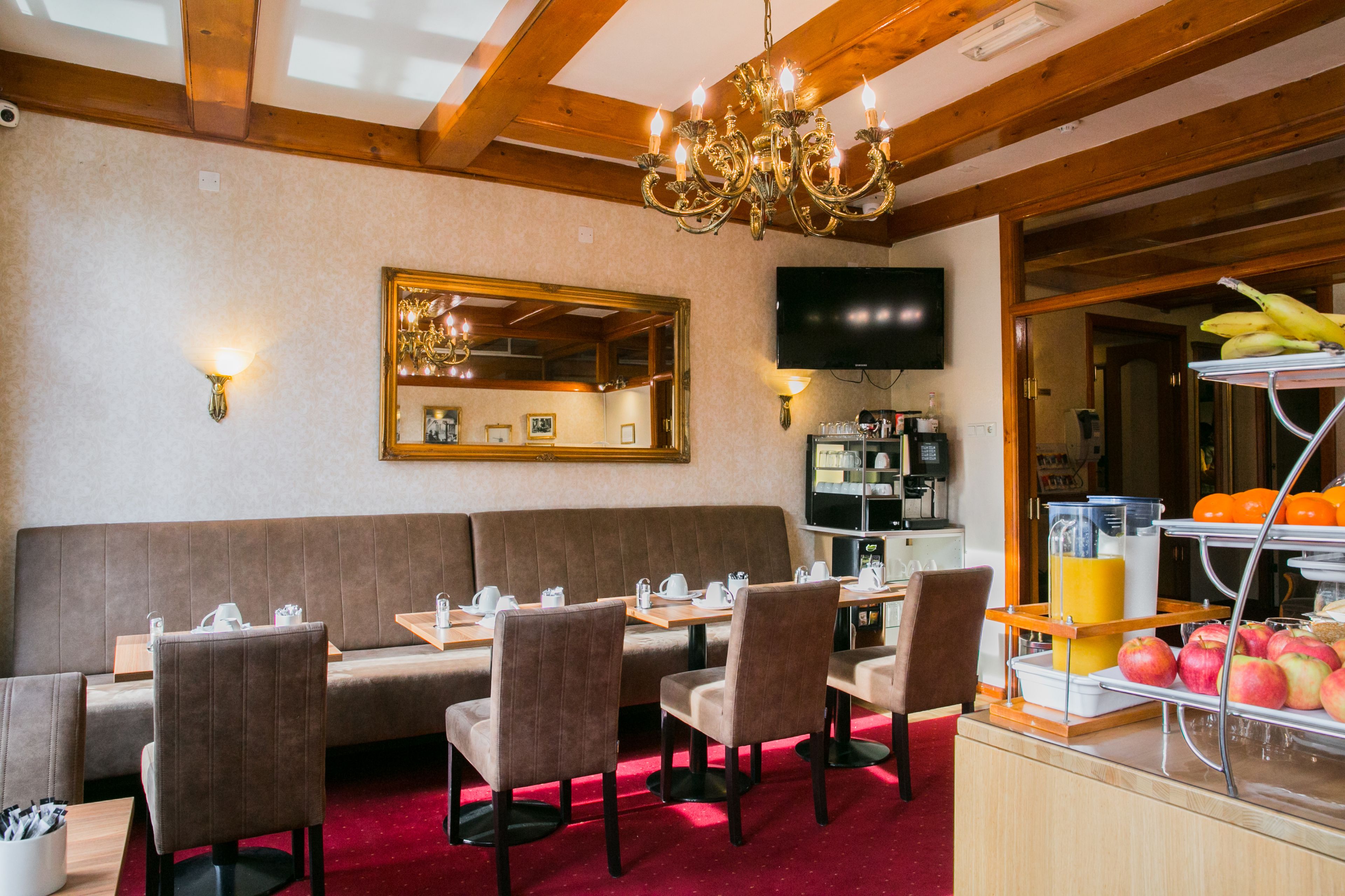 A restaurant with a wooden ceiling, a chandelier, a mirror, and a breakfast bar with fruits and beverages.