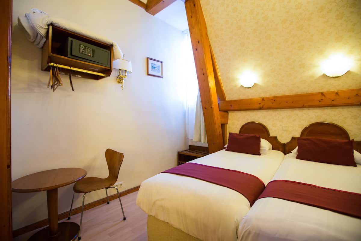 Habitación de hotel con dos camas, una silla, una mesita pequeña, una caja fuerte y una lámpara fijada a la pared.