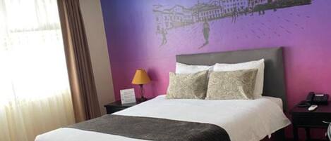 A hotel room with a bed, a chair, a nightstand, a lamp, and a wall mural.