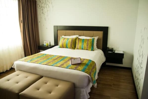 Quarto de hotel com cama grande, mesa de cabeceira, escrivaninha, um banco e janela com cortinas.