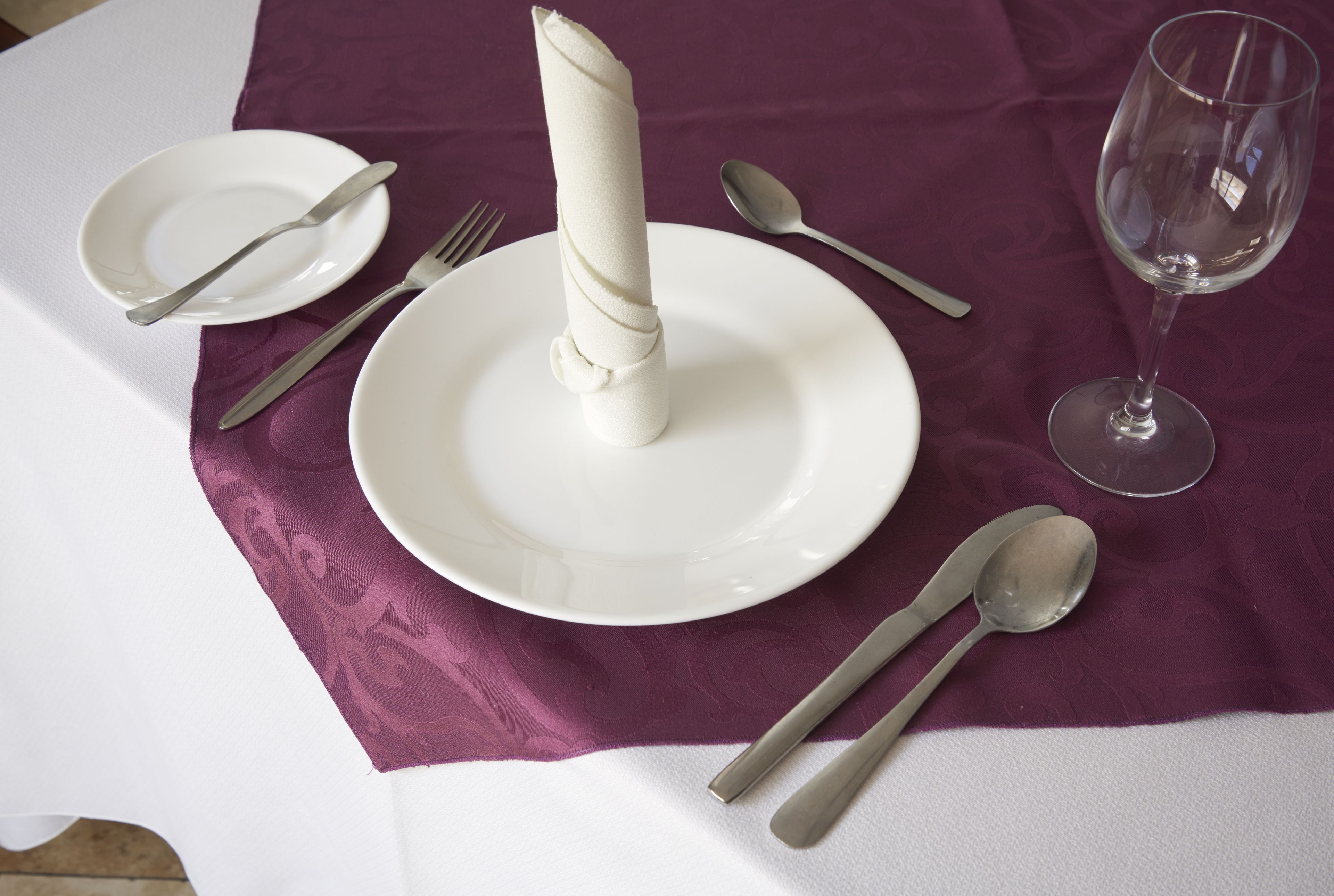 Ein Tisch ist mit weißen Tellern, Besteck und einem Weinglas auf einer violetten Tischdecke gedeckt.