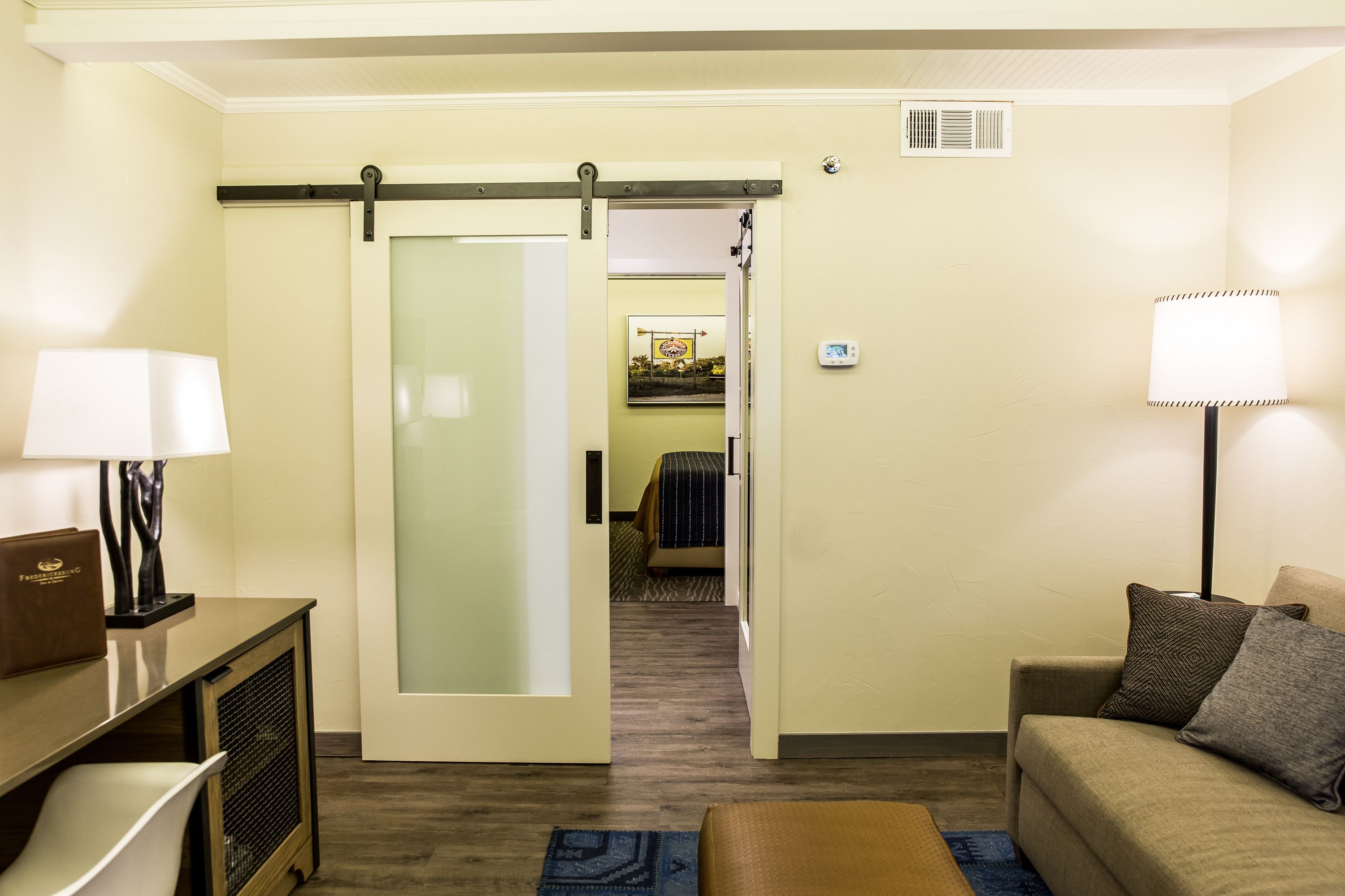 A hotel room with a sliding door leading to a bedroom, a sofa, a lamp, and a desk with a lamp.