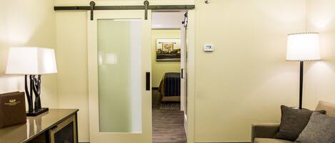 A hotel room with a sliding door leading to a bedroom, a sofa, a lamp, and a desk with a lamp.