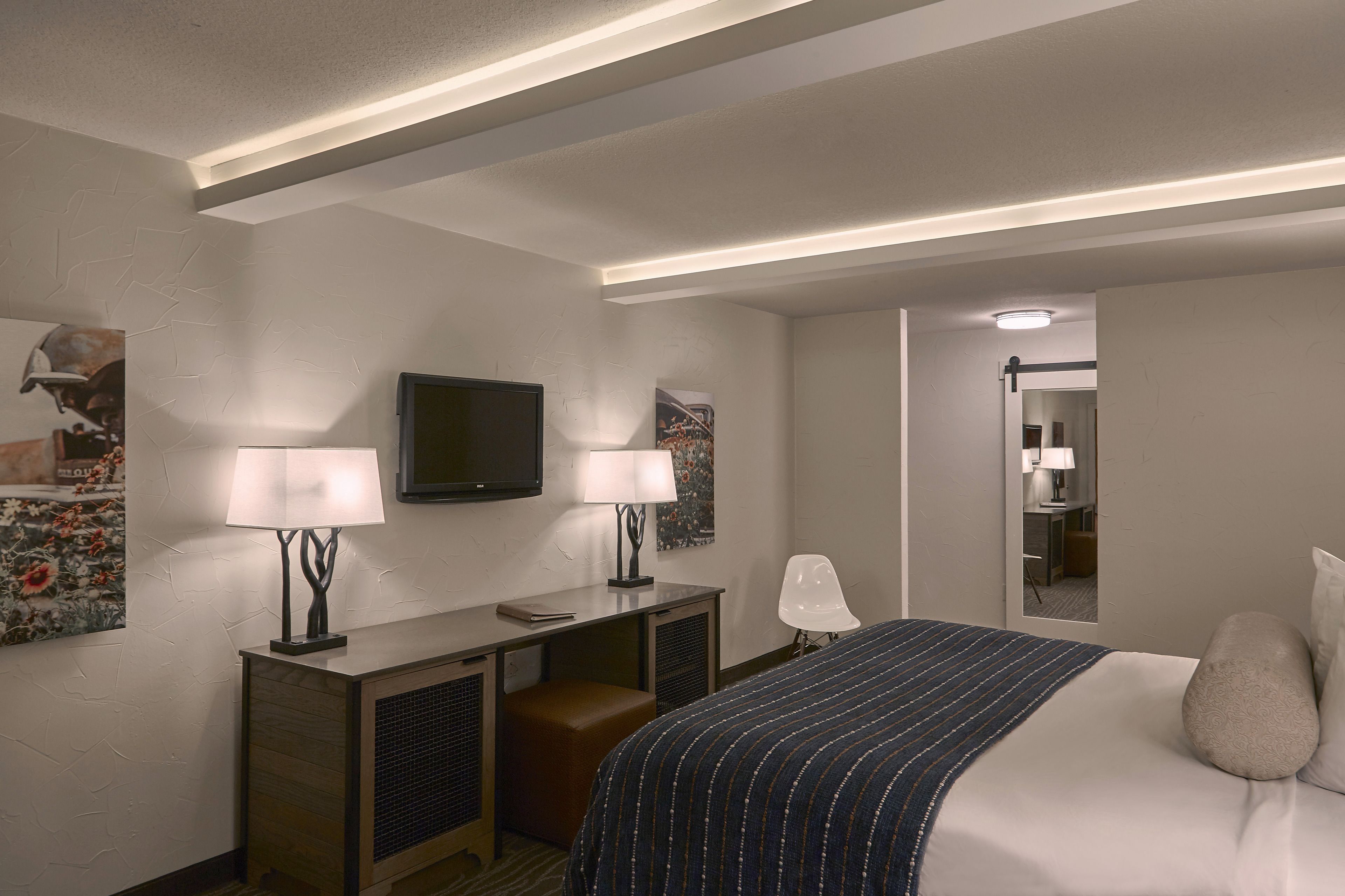 A hotel room with a bed, two bedside lamps, a flat-screen TV, a desk with a chair, and a small stool.