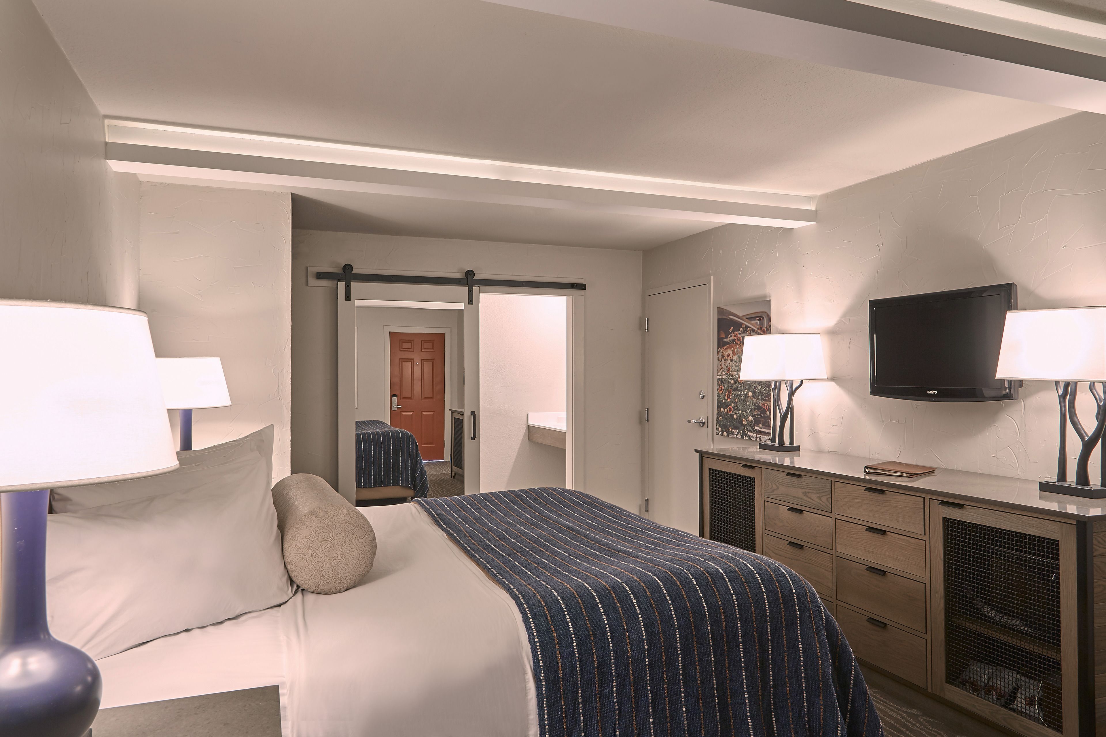 A hotel room with a bed, bedside lamps, a TV, a dresser, and a door leading to another room.