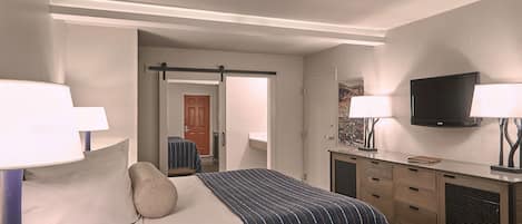 A hotel room with a bed, bedside lamps, a TV, a dresser, and a door leading to another room.