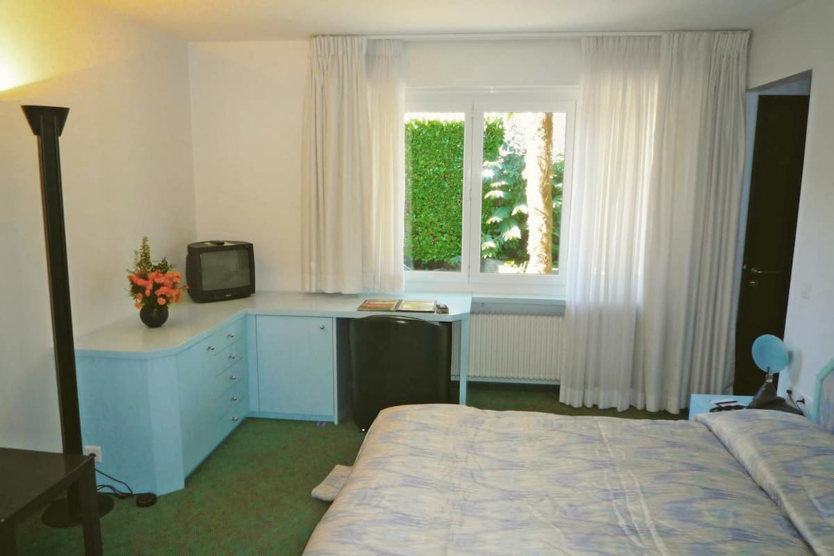 Un dormitorio con cama, escritorio, silla, televisor y ventana con cortinas.