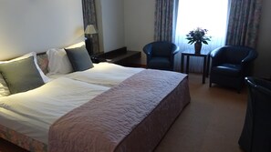 A hotel room with a large bed, two chairs, a small table, and a window with curtains.