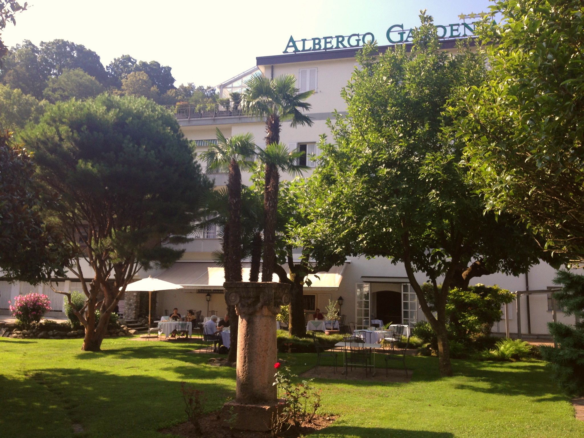 Un área de estar al aire libre con mesas y sillas, palmeras y un edificio con el letrero "Albergo Garden".