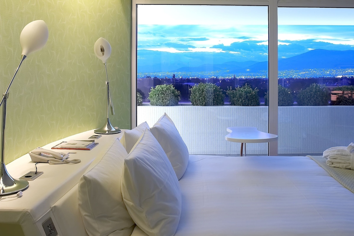 A hotel room with a bed, bedside lamps, a small table, and a view of a cityscape through large windows.