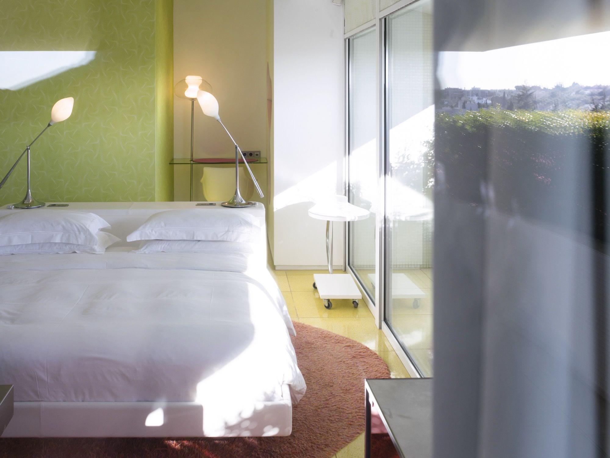 A modern hotel room with a large bed, two bedside lamps, a small table, and a view of the city through large windows.