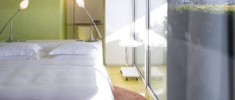 A modern hotel room with a large bed, two bedside lamps, a small table, and a view of the city through large windows.