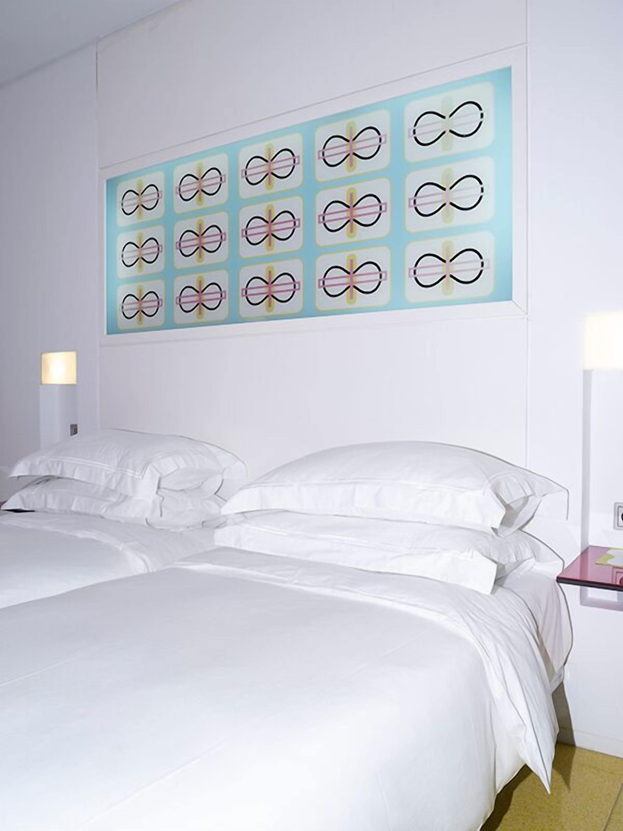 Une chambre d’hôtel avec un lit, du linge de lit blanc, une œuvre d’art accrochée au mur et des lampes de chevet.