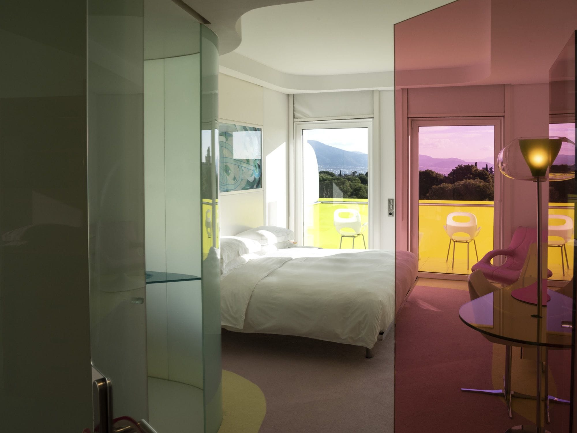 Un dormitorio moderno con cama, separador de vidrio, balcón con sillas amarillas y vista a las montañas.