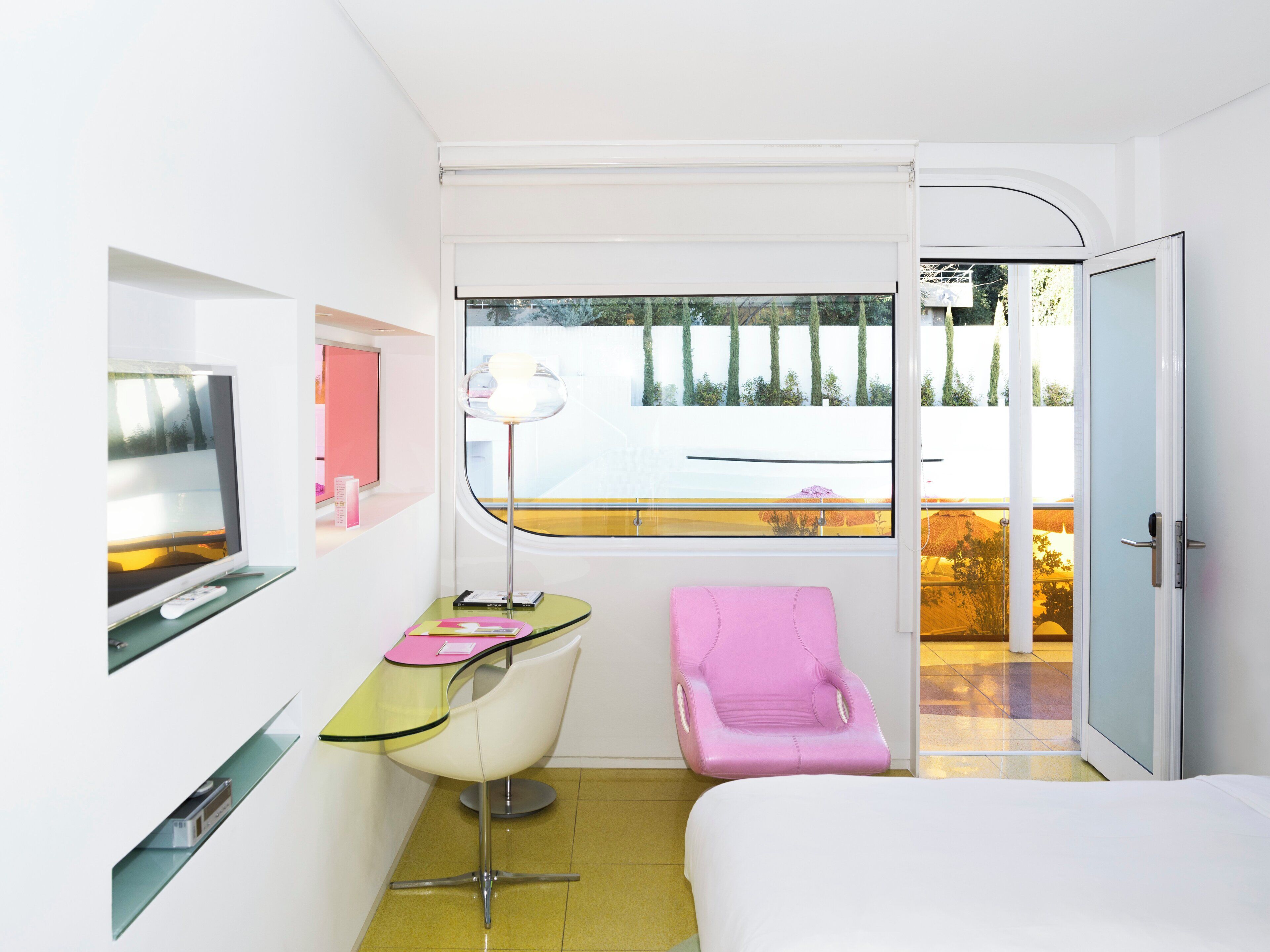 A modern hotel room with a bed, a desk with a chair, a massage chair, a TV, and a window with a view of trees and a building.
