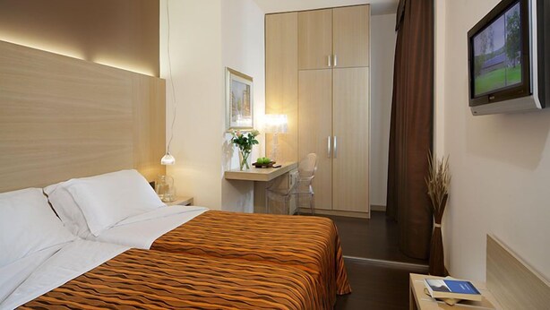 Ein Hotelzimmer mit Bett, Schreibtisch mit Stuhl, Kleiderschrank, Fernseher und einer Vase mit Trockenblumen.