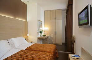 Habitación de hotel con cama, escritorio con silla, armario, televisor y un jarrón con plantas secas.