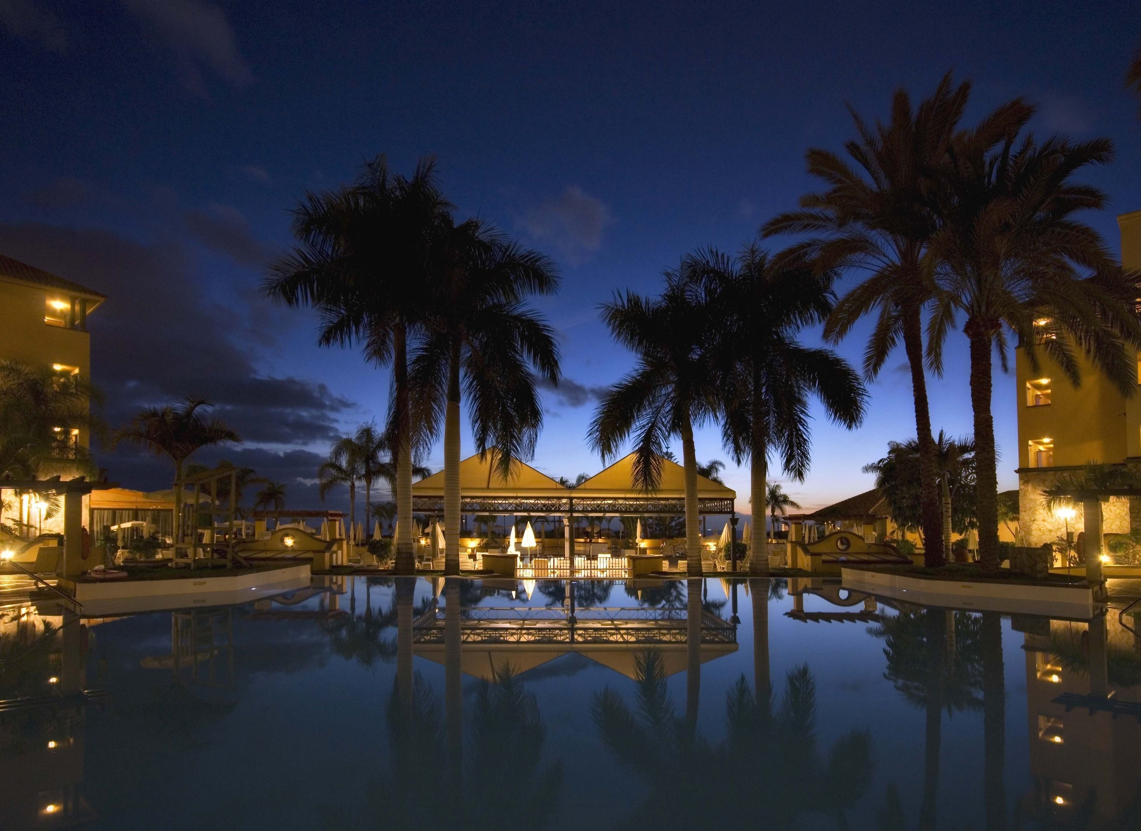 A hotel with a large reflective pool, palm trees, and a covered area with seating.