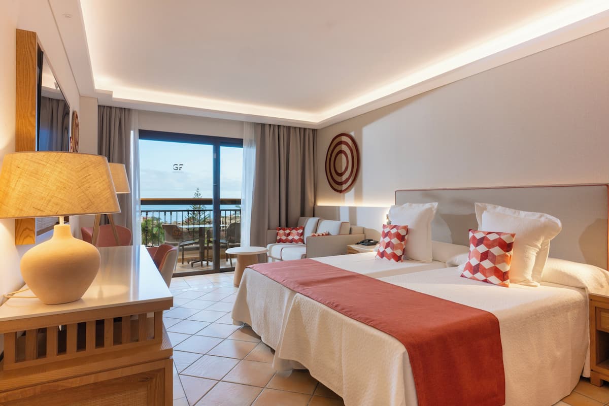 A hotel room with a bed, bedside tables, a desk with a lamp, a balcony with a view, and a target decoration on the wall.
