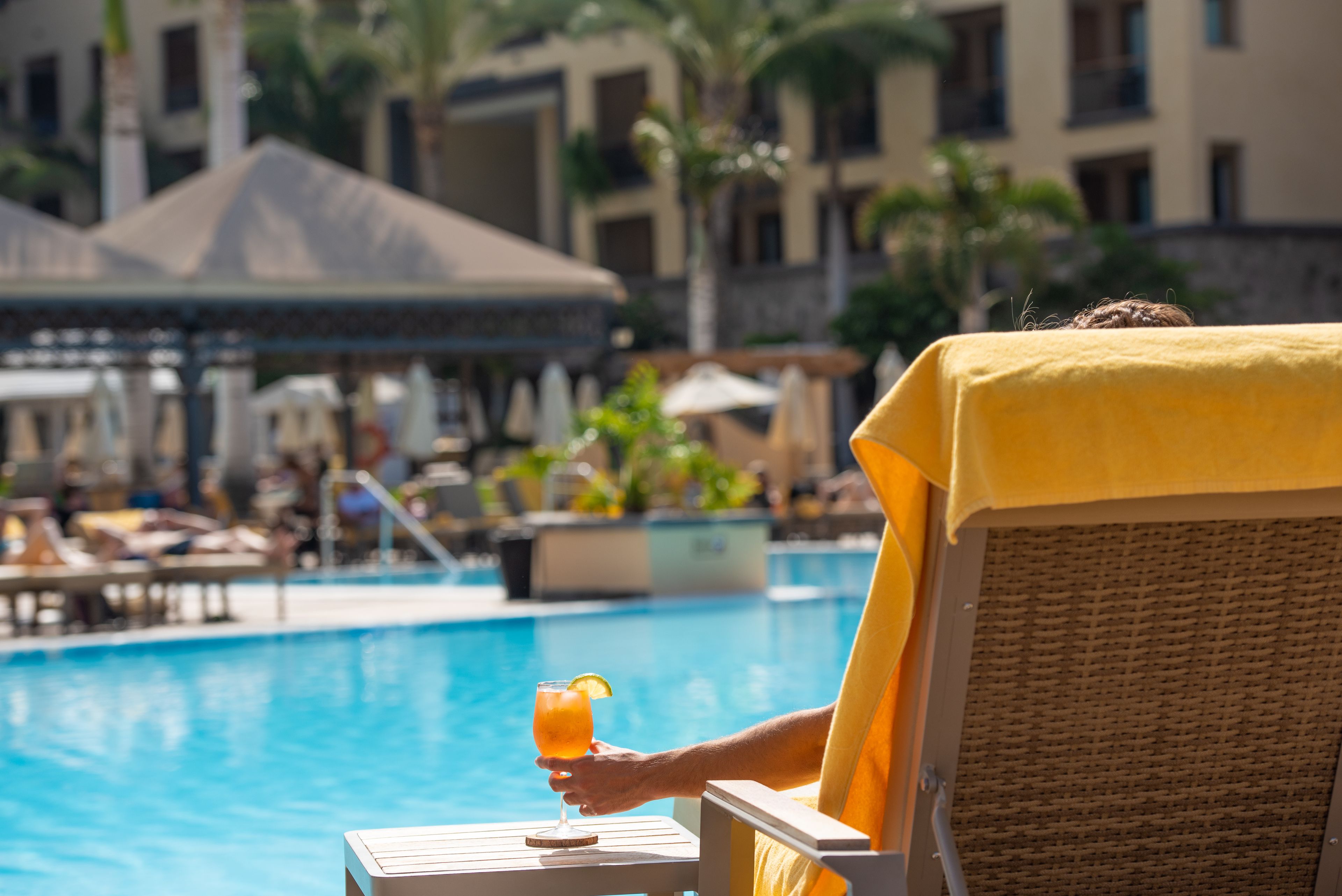 Une personne se détend près d’une piscine, assise sur une chaise longue et tenant un verre à la main, avec vue sur l’espace piscine, agrémenté de parasols et d’autres chaises longues.