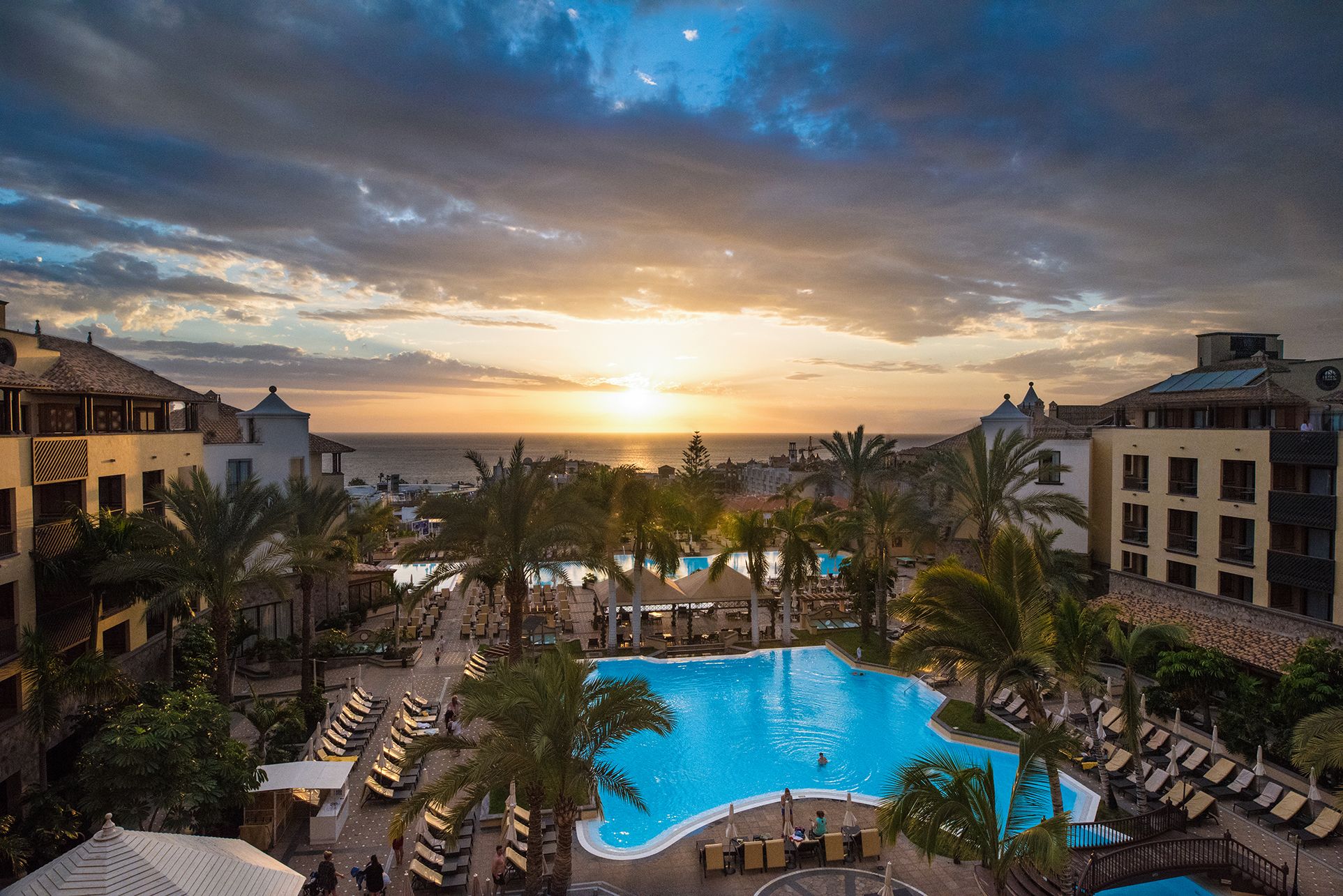 A resort with a large swimming pool surrounded by palm trees and lounge chairs, overlooking a beach and the ocean at sunset.