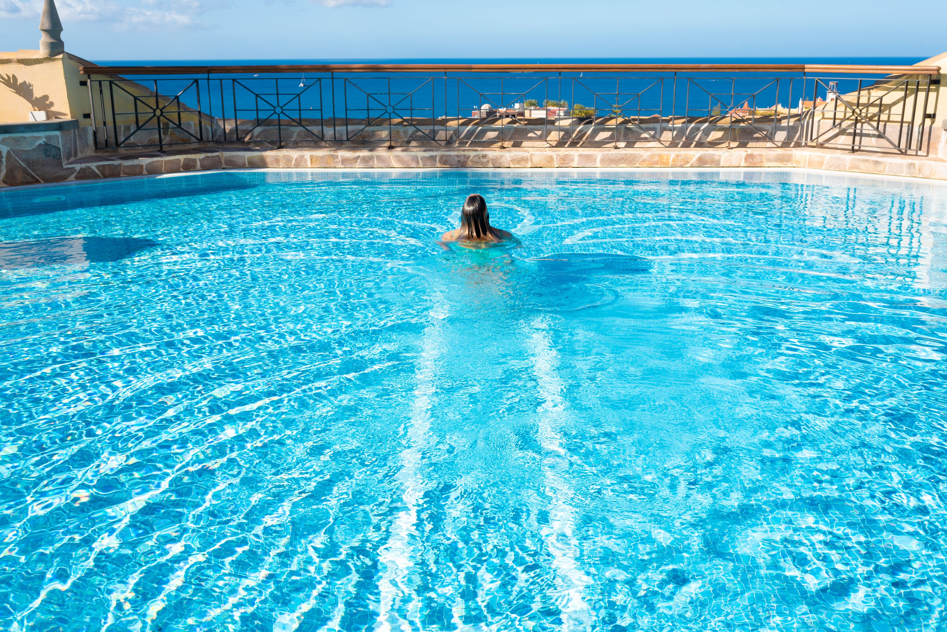 Una piscina azul cristalina con una persona nadando, rodeada por un muro de piedra y vistas al mar.