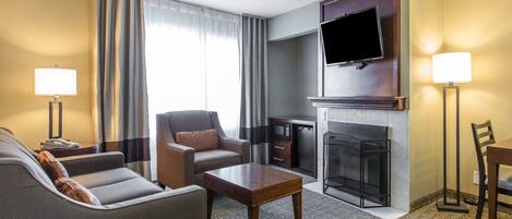 A hotel room with a sofa, armchair, coffee table, flat-screen TV, fireplace, and a dining area with a table and chairs.