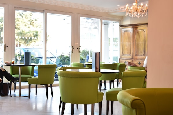 A room with green chairs and tables, large windows, and a chandelier.