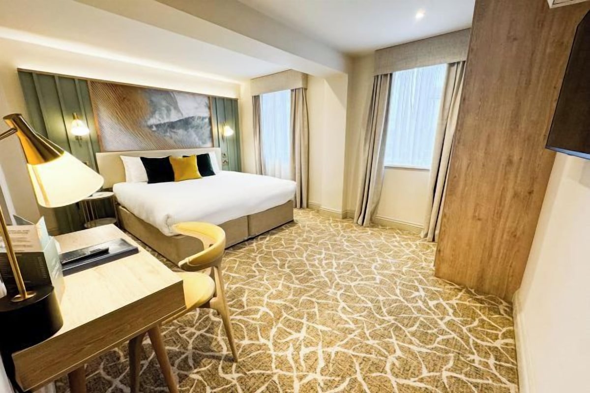 A hotel room with a bed, a desk with a lamp, a chair, and a large window with curtains.