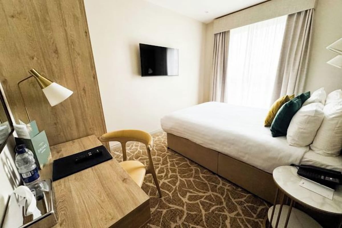 A hotel room with a bed, a desk with a lamp, a chair, a TV on the wall, and a small table with a remote control.