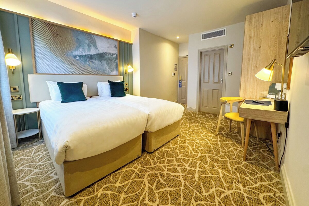A hotel room with a large bed, a desk, and a chair. There is a painting on the wall and a door in the background.