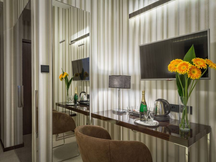 Ett modernt hotellrum med en stor spegel, en platt-TV, ett matbord i trä och en vas med blommor.