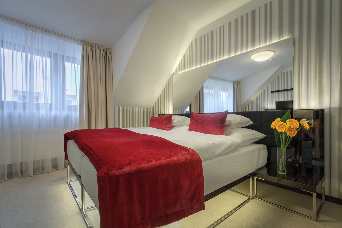 Quarto de hotel moderno com cama grande, roupa de cama vermelha, mesa de cabeceira com um vaso de flores e um espelho grande.